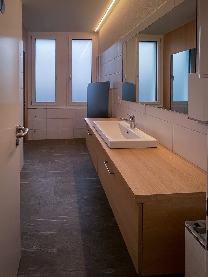 Spacious bathroom with wooden vanity countertop, white sink, tiled walls, and frosted glass windows. The room also features a private washing machine.