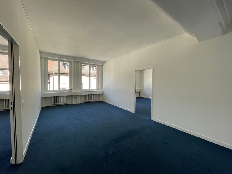 Large room with white walls, blue carpeted floor, and multiple windows providing natural light. The room appears to be empty, ready for furnishing and use.
