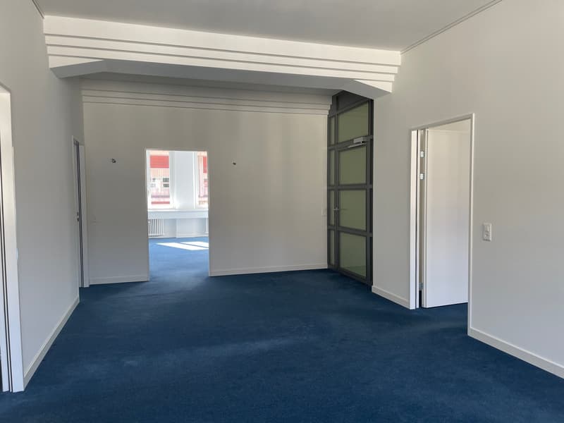 The image shows a large, open office space with gray walls, a blue carpeted floor, and a glass door leading to another room. The room has a modern, minimalist design with clean lines and a spacious layout.