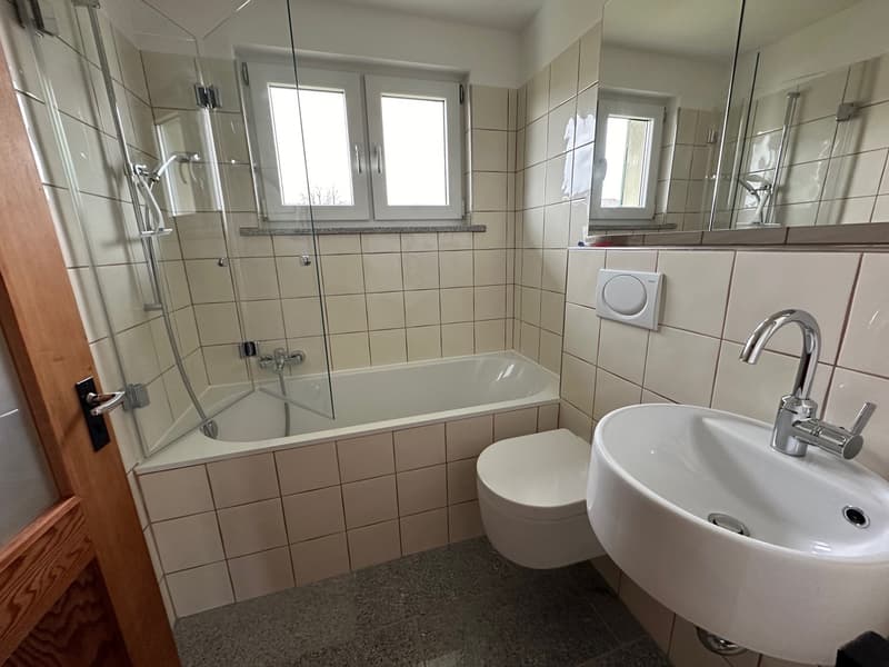 Spacious bathroom with tiled walls, a bathtub with glass shower enclosure, a vanity sink, and a toilet. The room has large windows providing natural light.
