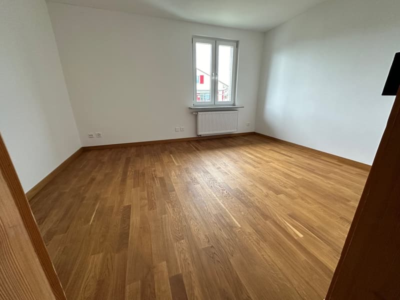 Spacious bedroom with hardwood flooring, white walls, and a large window providing natural light.