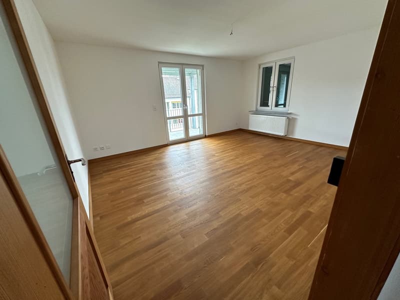 Spacious living room with hardwood flooring, white walls, and a sliding glass door leading to a balcony or terrace.
