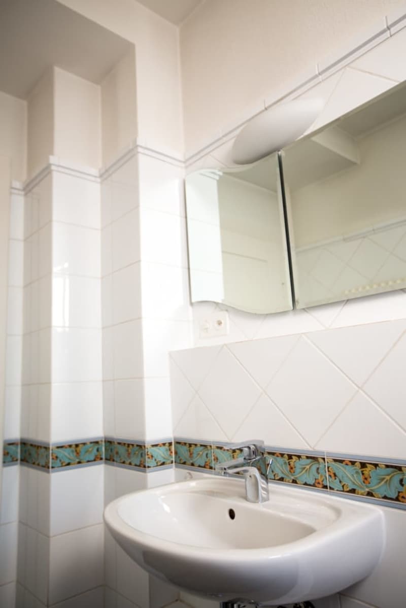 White tiled walls, white countertop, silver faucet, mirror, tiled borders
