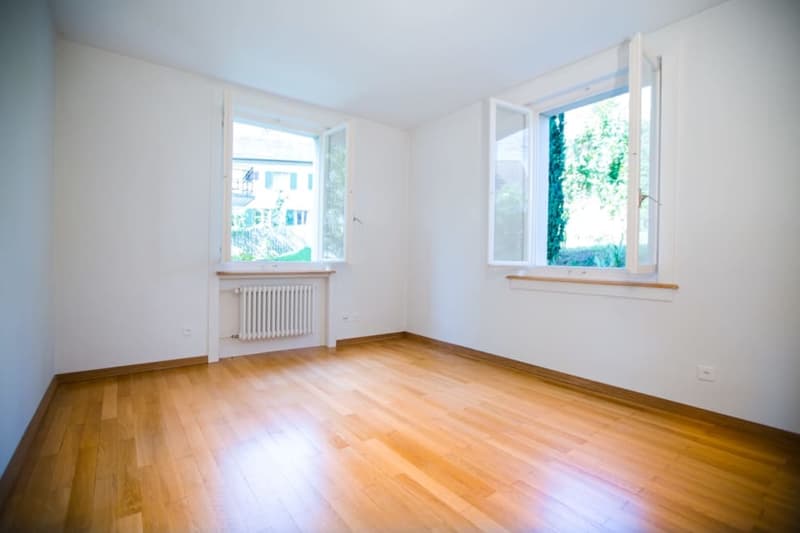 An empty room with hardwood floors, white walls, and large windows that offer a view of the outdoors.