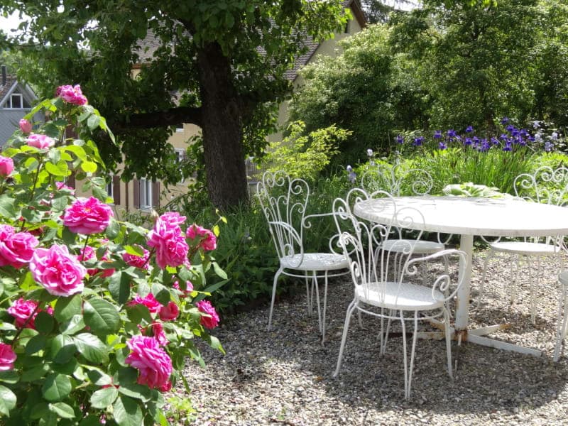 a small graveled garden area with a table and chairs, surrounded by pink flowers and trees, a house and other greenery in the background
