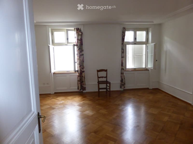 Spacious room with hardwood flooring, white walls, large windows, and a wooden chair in the corner.
