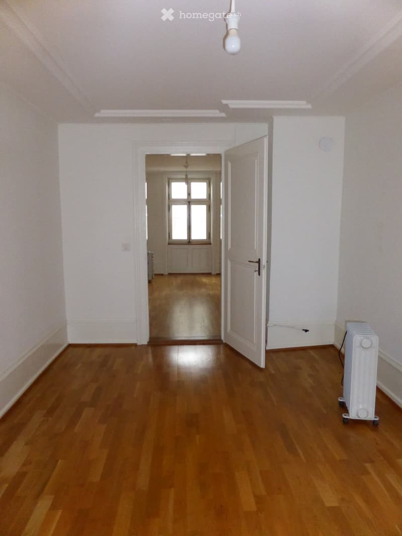 The image shows an empty room with white walls, a hardwood floor, and a single light bulb hanging from the ceiling. There is a door leading to another room visible in the background.