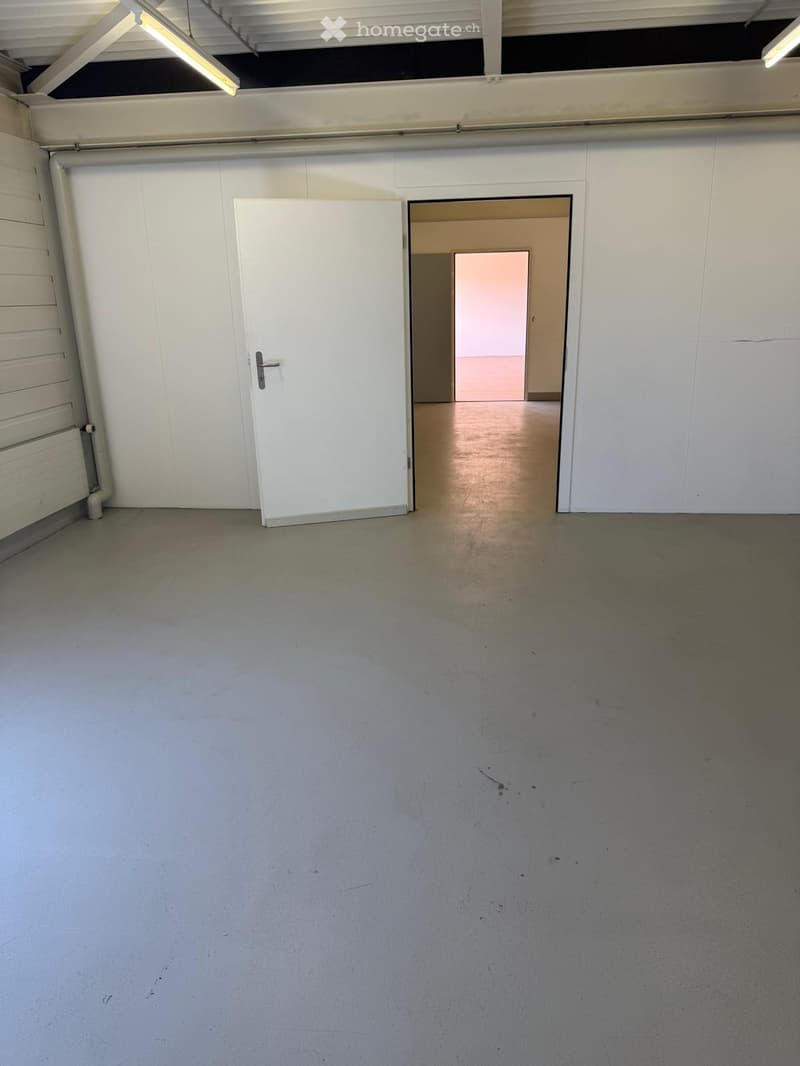 The image shows a large, open room with white walls and a concrete floor. There is a white door on the left side of the room, and an open doorway leading to another room on the right side. The ceiling has exposed beams and lighting fixtures. The room appe