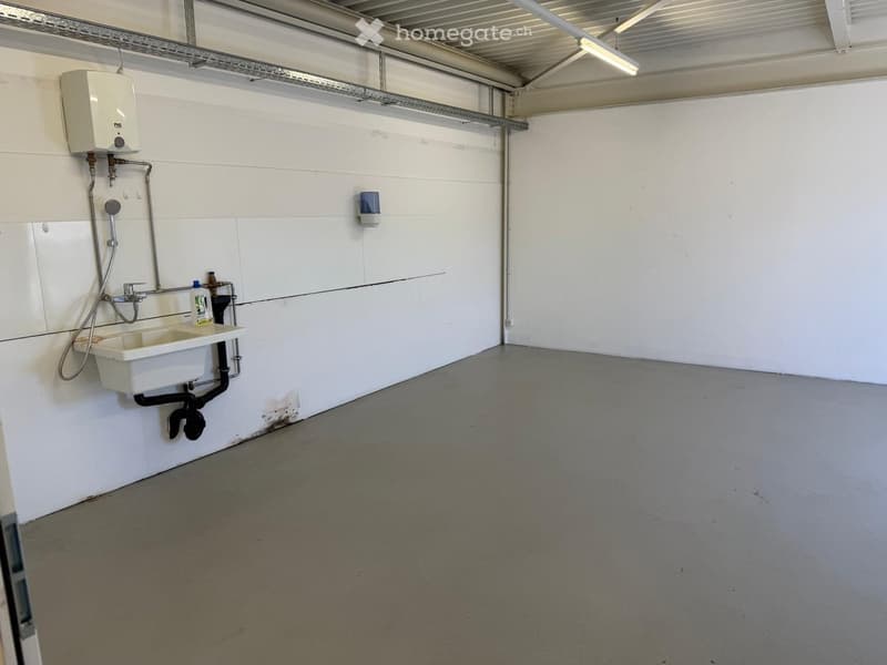 This image shows the interior of a garage. The space has a concrete floor, white walls, and a high ceiling with exposed beams. There is a utility sink and plumbing fixtures installed on one wall, indicating this garage may have additional uses beyond just