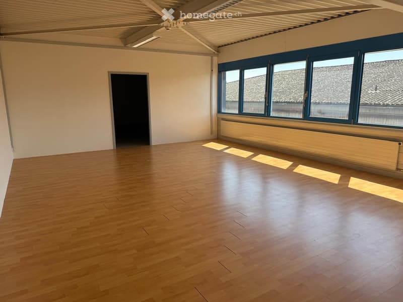 Large open room with hardwood floors, high ceilings with exposed beams, and large windows providing natural light. The room appears to be empty, ready for furnishing and use.