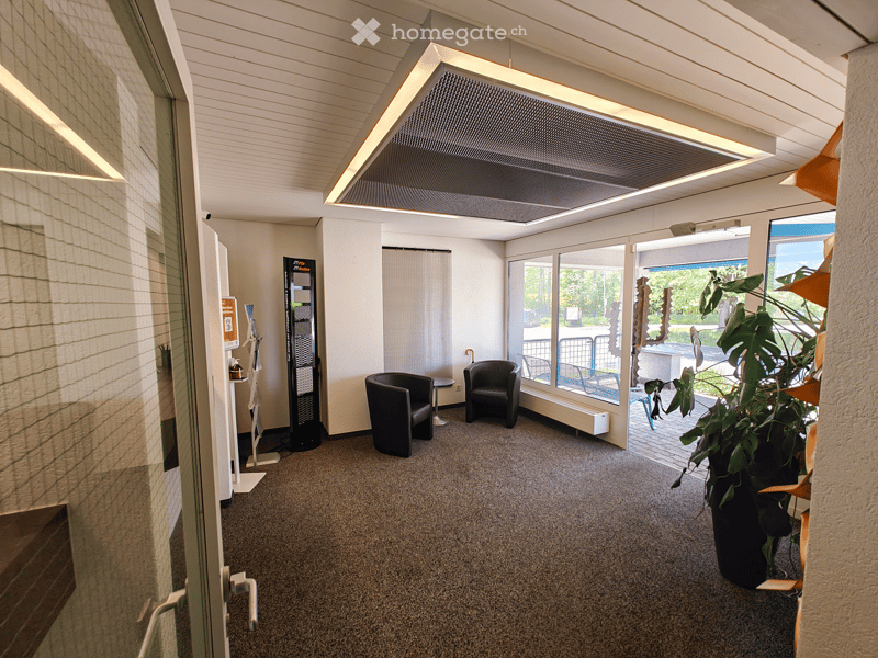 Spacious office space with large windows, carpeted floor, two black armchairs, and a vending machine. The room has a modern, open design with a suspended lighting fixture.