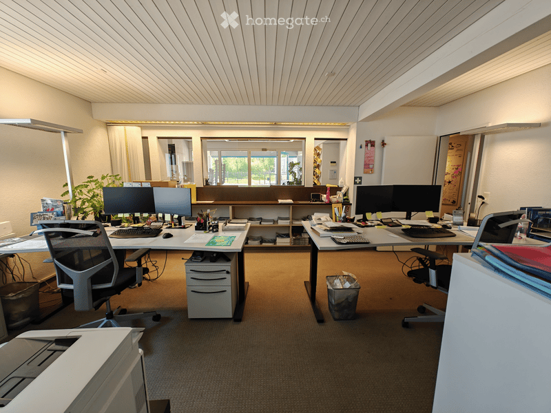 The image shows a cluttered office space with multiple desks, computers, and office equipment. The room has a high ceiling with a paneled wooden design. There are large windows providing natural light, and the overall atmosphere appears to be a profession