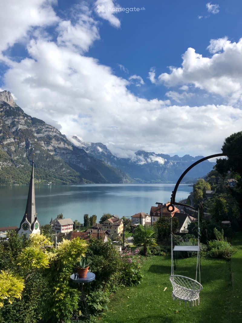 A garden with a swing chair and plants, overlooking a lake and mountains