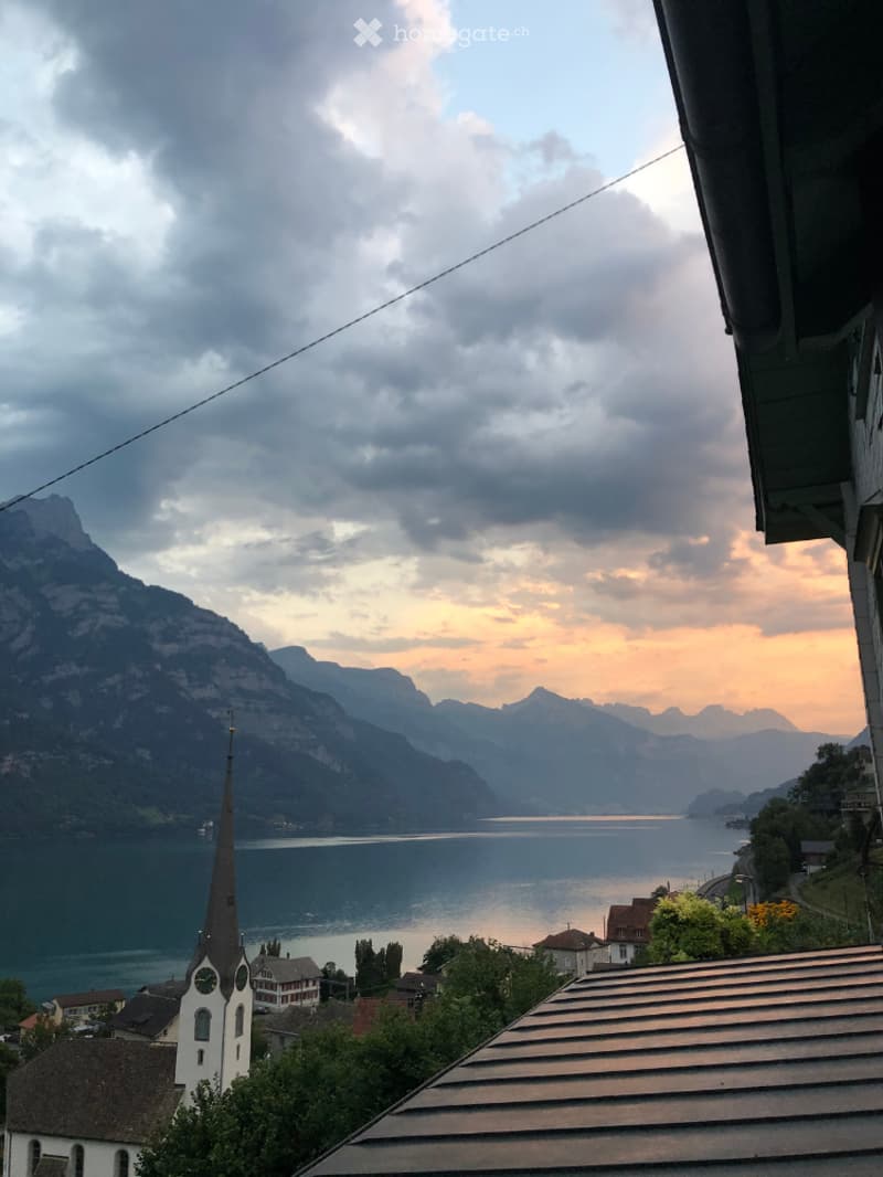 Scenic view of a lake surrounded by mountains, with a village and church spires in the foreground. The sky is filled with dramatic clouds, creating a beautiful sunset over the landscape.