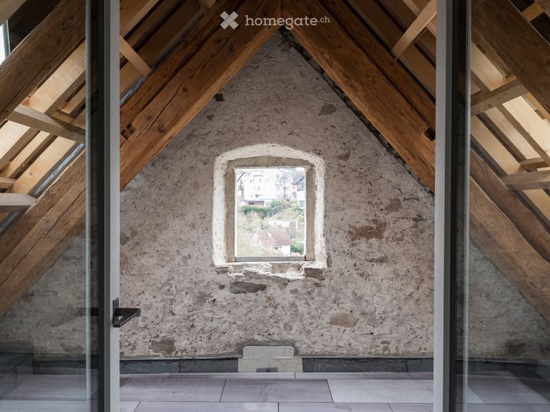 An attic room with sloping ceilings, exposed wooden beams, a small window, and a tiled floor.