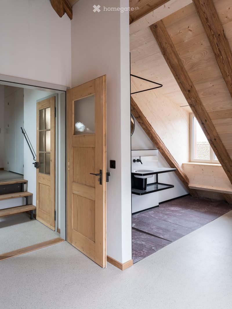 a small room with wooden door, a sink with mirror and wall-mounted wooden shelves, sloped wooden ceiling, wooden windows and flooring, open staircase