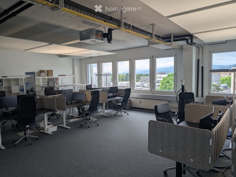 Open office layout with desks, chairs, and computer equipment. Large windows providing natural light and a view of the city outside. Suspended ceiling with lighting fixtures. Shelving units and storage areas along the walls.