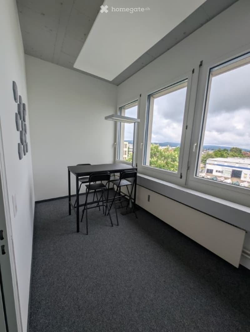 The image shows a small office space with a table and chairs, large windows providing natural light, and a concrete ceiling. The room has a minimalist, modern design with gray walls and dark carpeting.