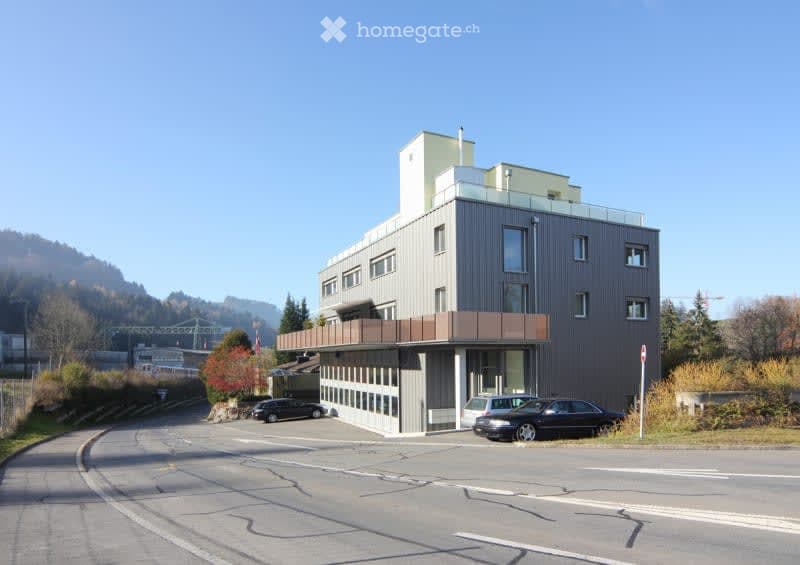 Multi-story commercial or office building with a modern, angular design. The building has a gray exterior with large windows and a raised ground floor. There is a parking area in front of the building.