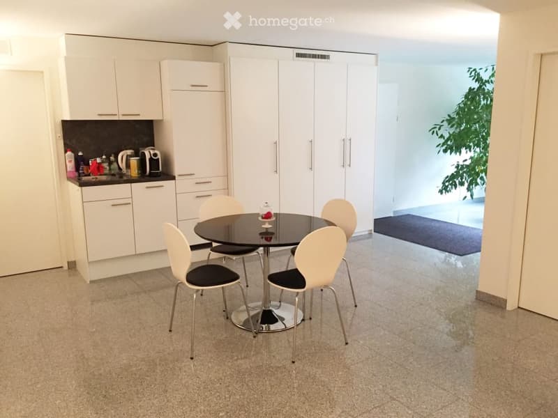 The image shows a modern kitchen and dining area. The kitchen features white cabinets and a black countertop. There is a dishwasher visible in the kitchen. The dining area has a round glass table with four white chairs. The room has a bright and open feel