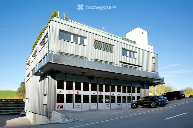 Modern multi-story commercial building with a gray metal facade, large windows, and a balcony on the upper floor. The building has the name 'Ichick motor' displayed on the front. There are several cars parked in front of the building.