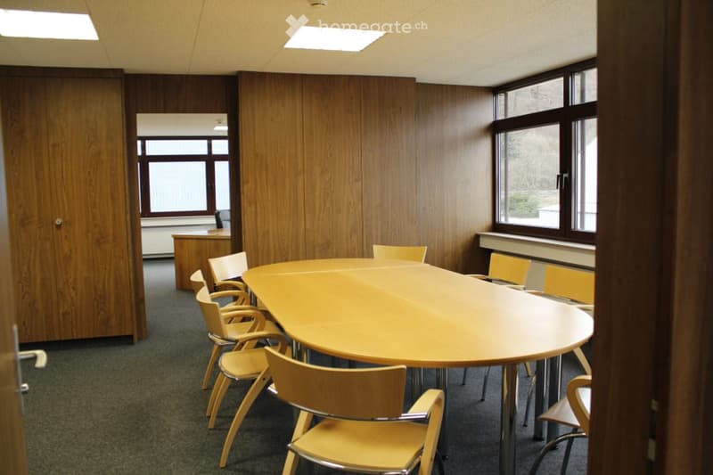 Large oval wooden table with yellow chairs, wooden paneled walls, large windows, and recessed lighting in the ceiling.