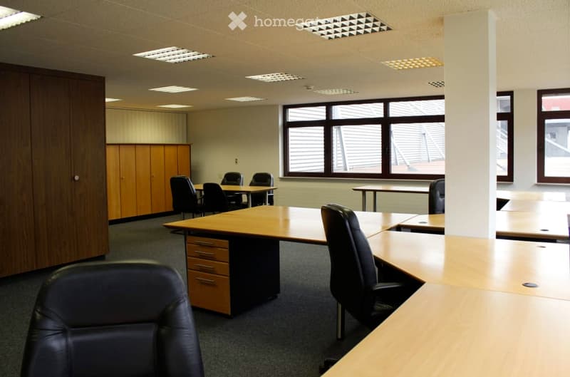 Large open office space with wooden desks, black leather chairs, and storage cabinets. The room has large windows providing natural light and recessed lighting fixtures on the ceiling.