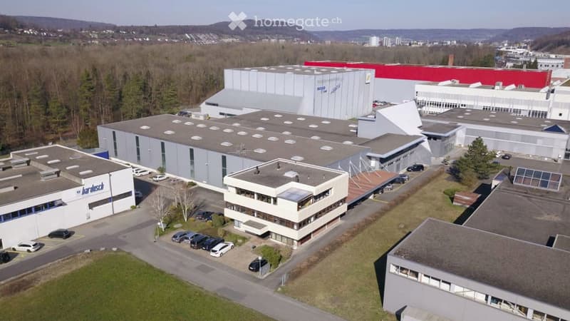 Large commercial building with a flat roof, white and gray exterior, and a parking lot in the foreground. The building appears to be a factory or warehouse based on its size and industrial design.
