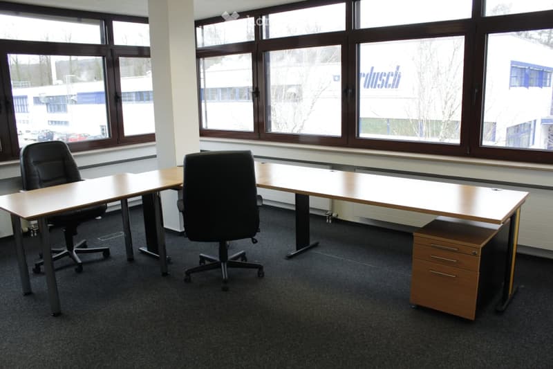 The image shows an office space with a large desk, two office chairs, and a filing cabinet. The room has large windows overlooking an outdoor area. The walls are plain and the floor is carpeted.