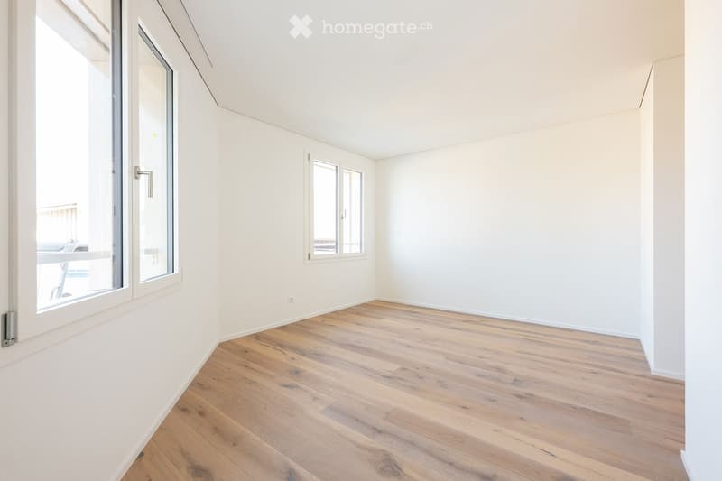 Empty room with light wooden flooring and white walls, featuring large windows and a door leading to a balcony.