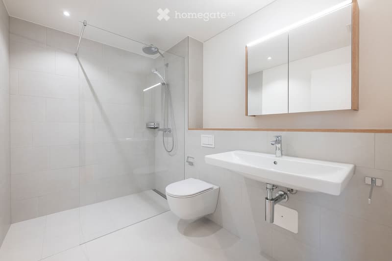Modern bathroom with white tiles, a glass-enclosed shower, a toilet, a sink with a mirror, and built-in lighting above the mirror.