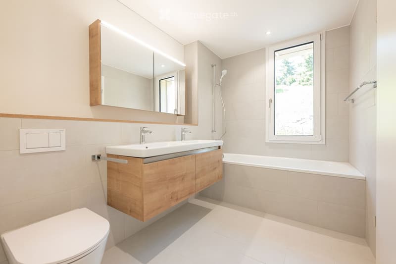 Modern bathroom with a double sink, toilet, bathtub, large mirror, and a window offering a view of greenery.