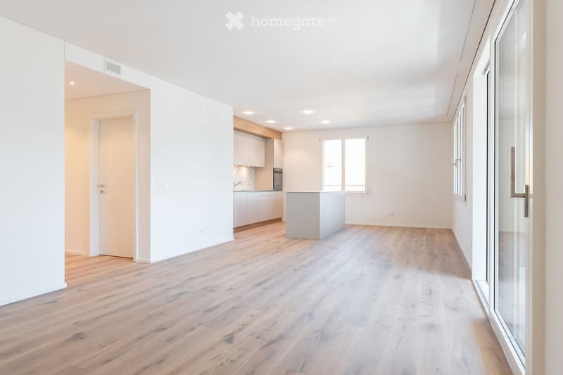 Bright, open living space with light wooden flooring, white walls, and a modern kitchen area featuring cabinets and a countertop.