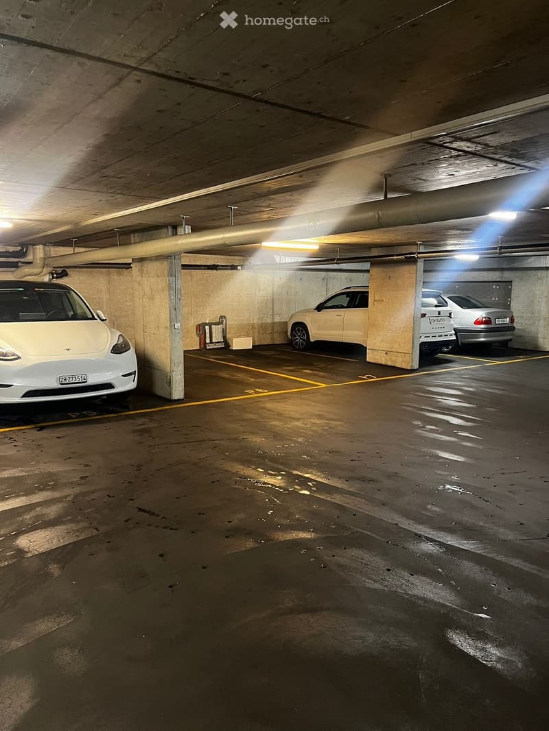 Underground parking garage with concrete walls and ceiling, several parked cars including a Tesla Model 3 and another vehicle, wet floor indicating recent cleaning or flooding