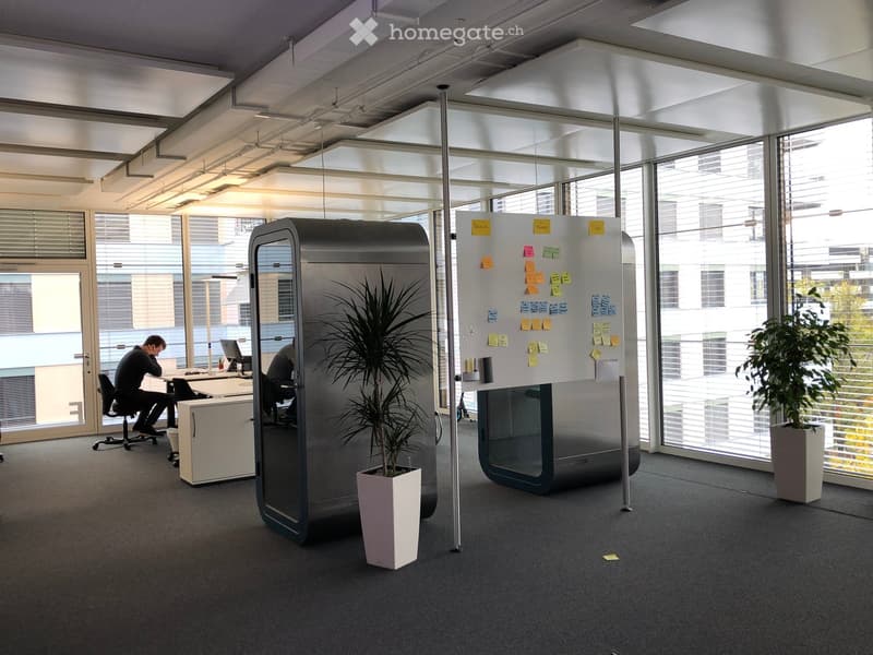 The image shows an open office space with large windows, a whiteboard with sticky notes, a refrigerator, and a potted plant. There are two people working at desks in the background.