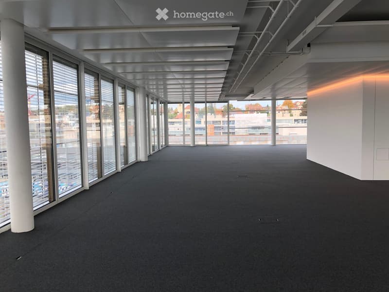 Large open-plan office space with high ceilings, exposed beams, and floor-to-ceiling windows offering a panoramic view of the surrounding cityscape. The space features a grey carpeted floor and a clean, modern aesthetic.
