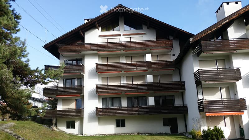 3-story apartment building with white exterior walls, wooden balconies, and a slanted roof. The building is surrounded by pine trees and has a landscaped garden area in the front.