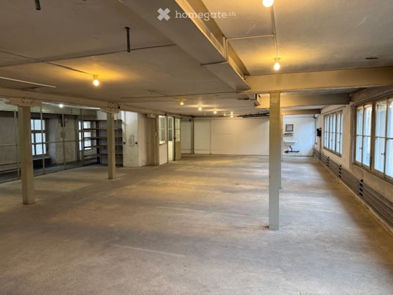 large room, exposed beams, concrete floor, metal columns, multiple large windows, some with grills