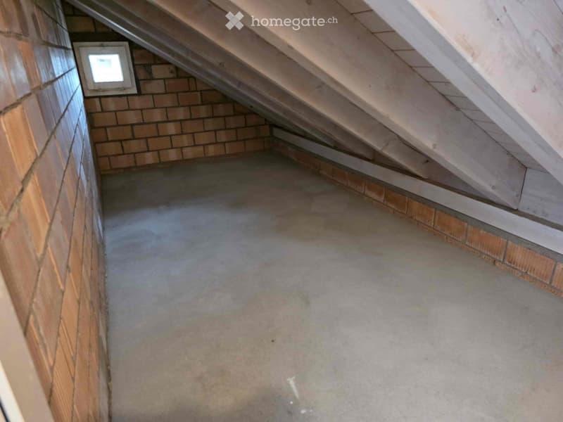 The image shows an attic space with exposed wooden beams and brick walls. The floor appears to be a concrete slab. There is a small window visible in the wall, providing some natural light to the space.