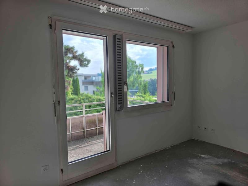 The image shows a room with large windows that provide a view of the outdoor greenery. The room appears to have a sliding glass door leading to a balcony or terrace area.