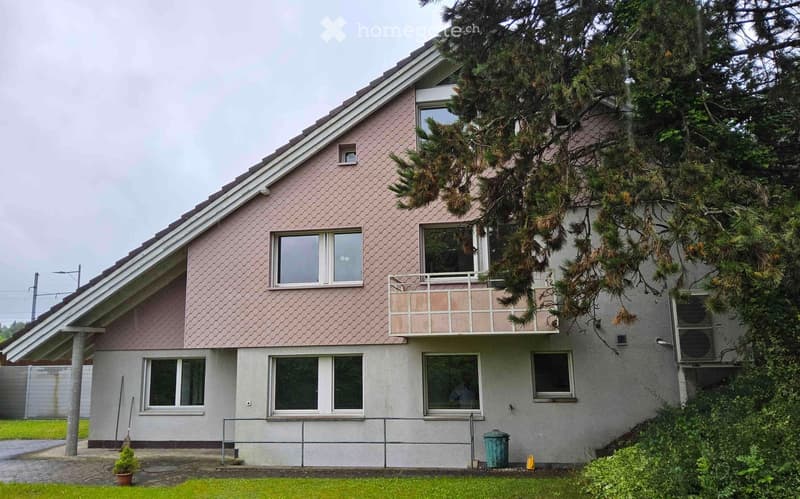 2-story house with a slanted roof, gray exterior walls, and large windows. There is a balcony or terrace on the upper level. The house is surrounded by trees and greenery.
