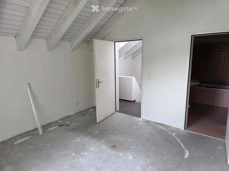 This image shows an unfinished interior space with white walls, a vaulted ceiling with exposed beams, and a concrete floor. There is an open doorway leading to another room and a built-in cabinet or shelving unit visible in the corner.