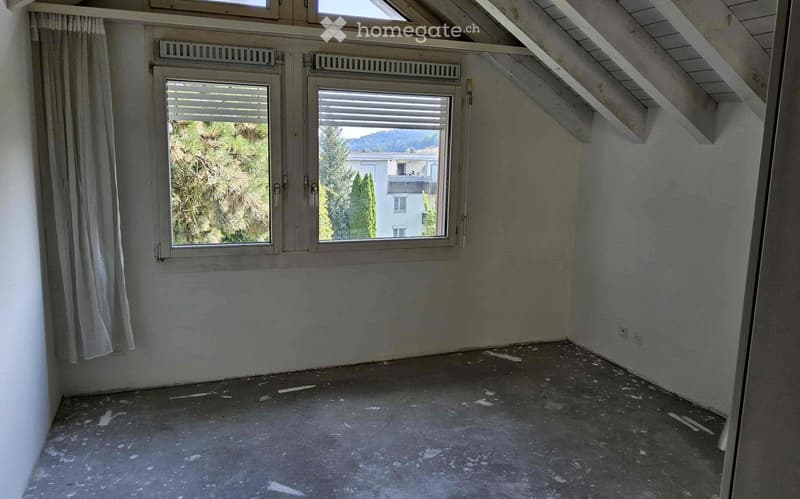 Attic bedroom with slanted ceiling, large windows providing a view of the surrounding trees and buildings, hardwood floor, and curtains on the windows.
