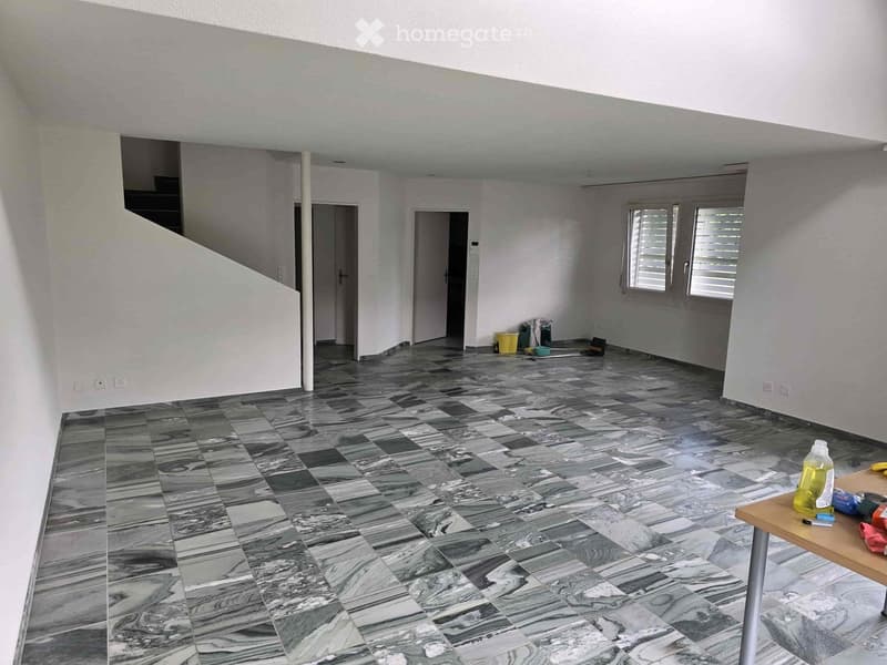 The image shows a large open space with a tiled floor in a gray and white marble pattern. The room has white walls and appears to be in the process of renovation or remodeling, with some cleaning supplies and tools visible on the floor. There are two door
