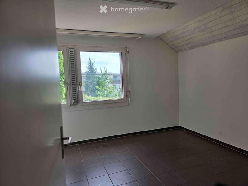 The image shows a room with white walls, a tiled floor, and a window overlooking some trees outside. The room appears to be empty, with no furniture or other items visible.