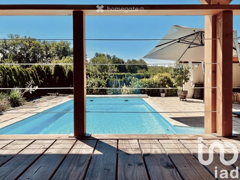 Outdoor patio with a wooden deck, a large swimming pool, and a white umbrella. The patio overlooks a lush, green landscape with trees and shrubs.