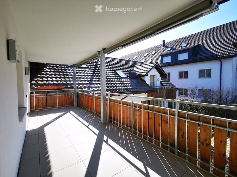 Spacious balcony with wooden railings, overlooking a residential neighborhood with tiled roofs and windows.