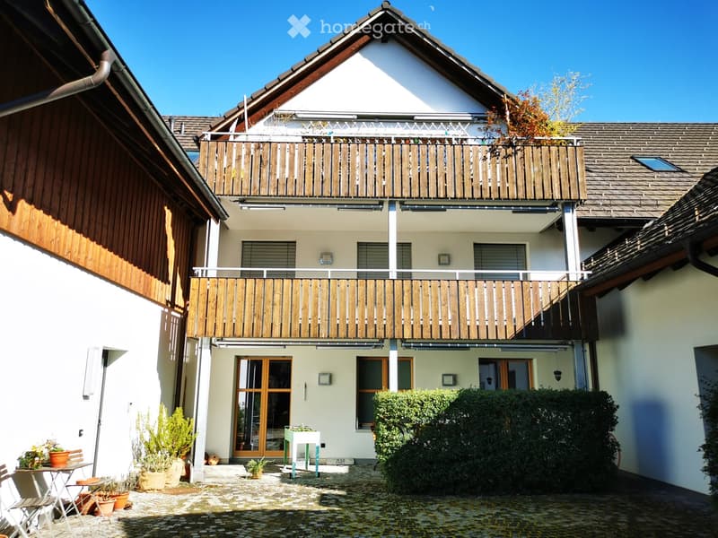 2-story apartment building with wooden balconies, white exterior walls, and a tiled roof. The building has several windows and a small courtyard or garden area in the front.