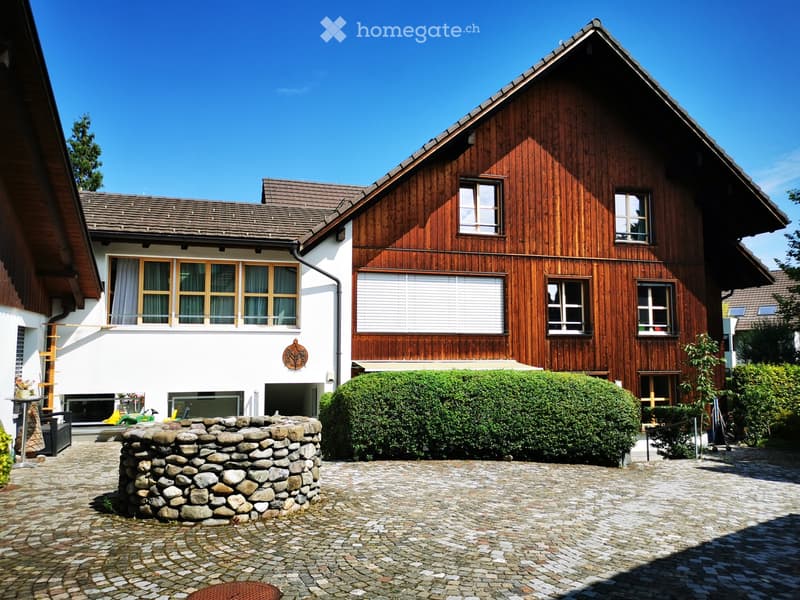 Two-story wooden house with white walls, large windows, and a garage. The house has a cobblestone driveway and a stone well in the front yard. The surrounding landscape includes trees and shrubs.
