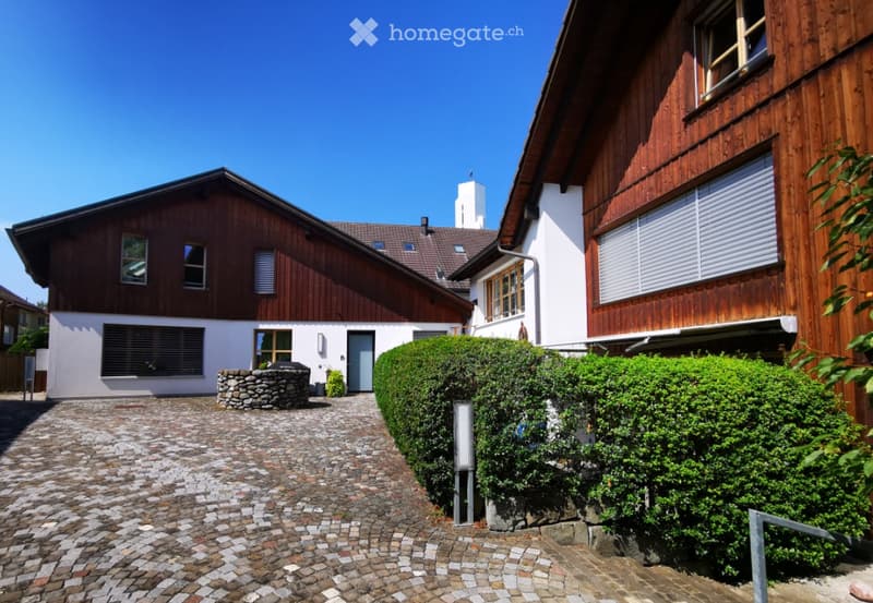 Two-story wooden house with a cobblestone driveway, surrounded by well-maintained hedges and landscaping. The house has a mix of wooden and white exterior walls, with several windows and a chimney visible.